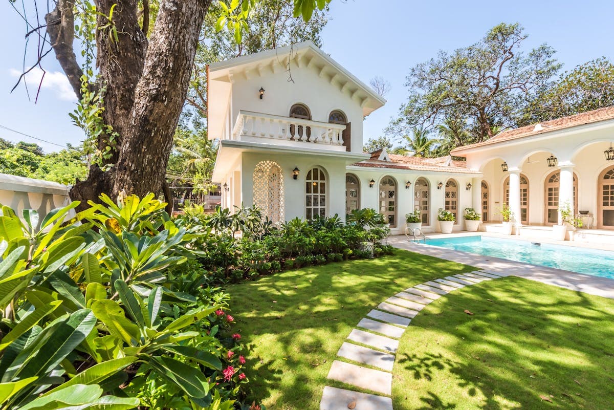 White colonial-style villa with an arched balcony and a pool, seen across a curving garden path