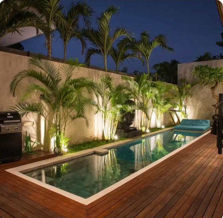 Lit lap pool at dusk on a timber deck, with uplit palms against a rendered wall, a barbecue and a turquoise sun lounger.