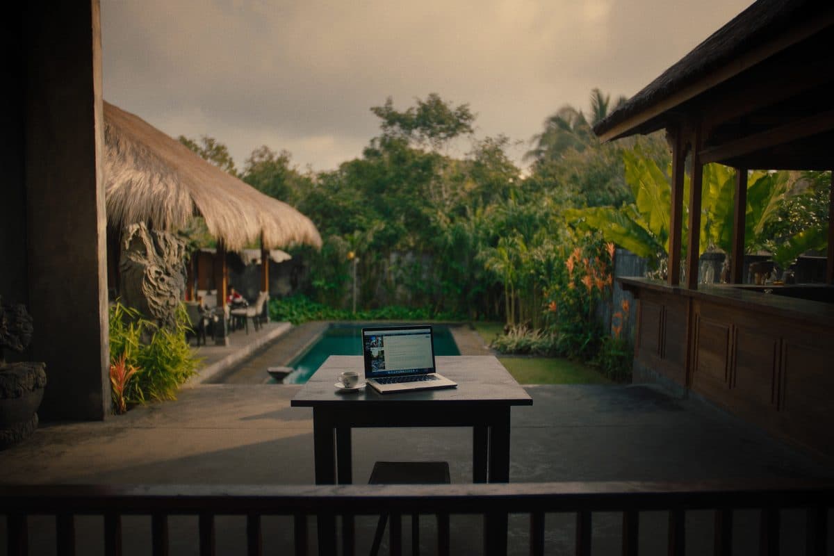 Open laptop and a coffee cup on a table on a Balinese villa terrace, overlooking the pool and garden