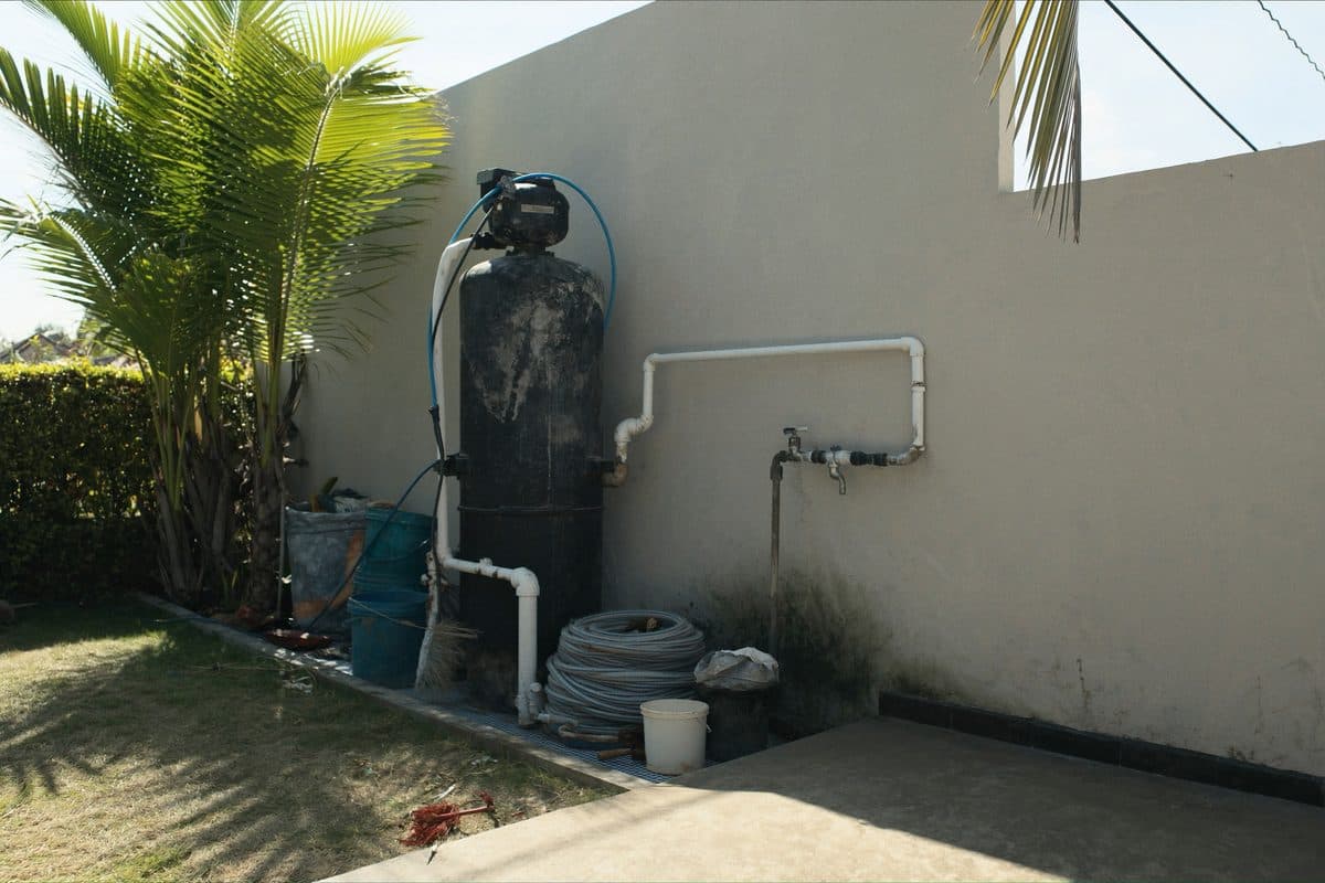 Water pump and filter tank plumbed against an exterior villa wall, beside a coiled hose