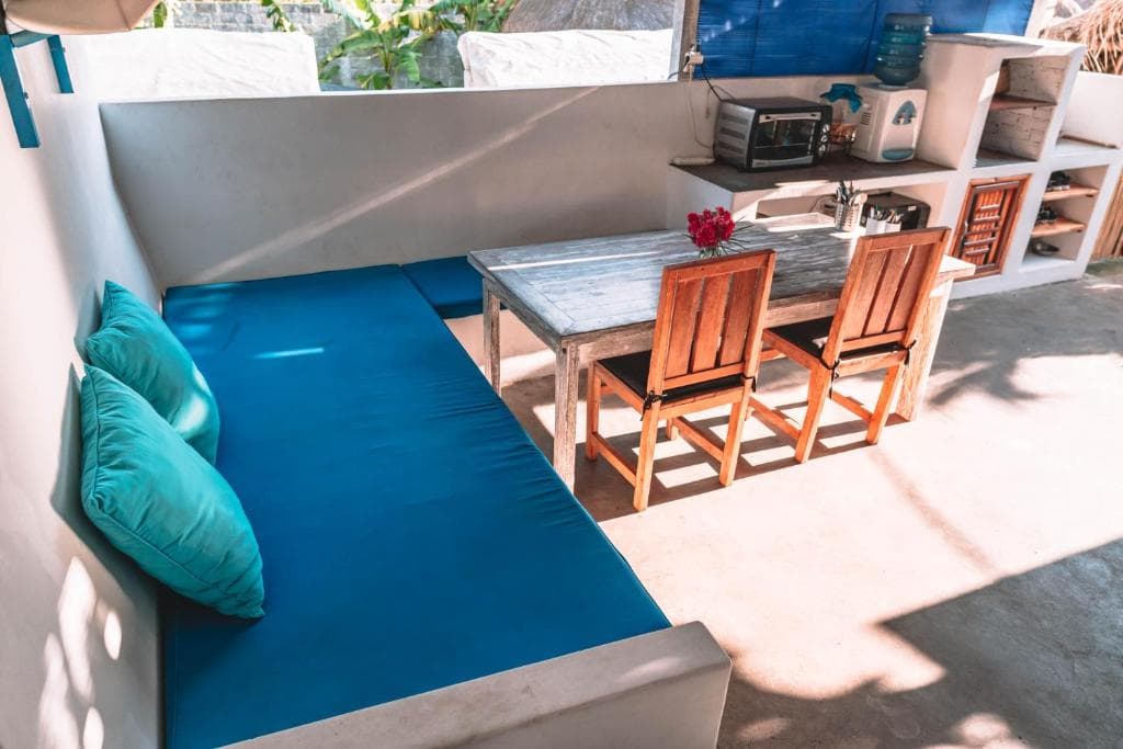 Open-air terrace with a built-in bench and turquoise cushions, a weathered wooden table and chairs, and a simple outdoor kitchen.