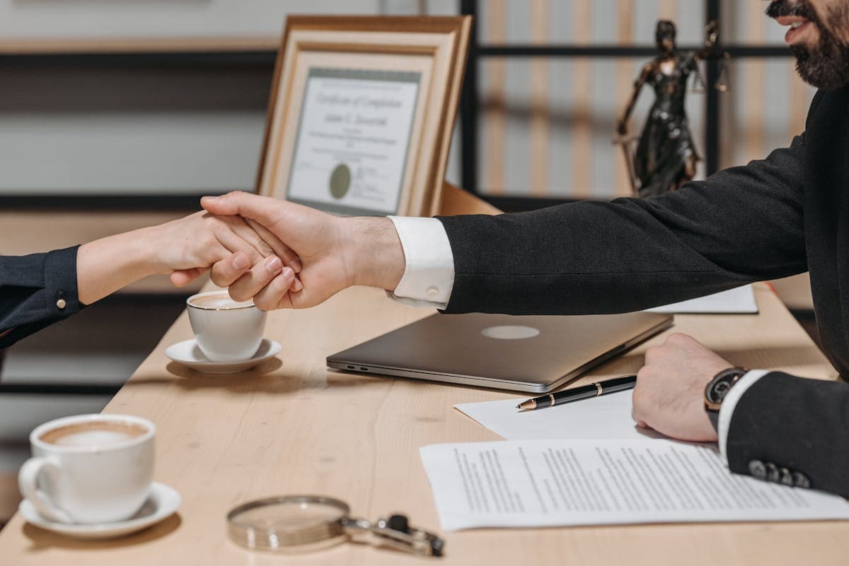 Two people in business dress shaking hands across a desk of contracts and coffee cups