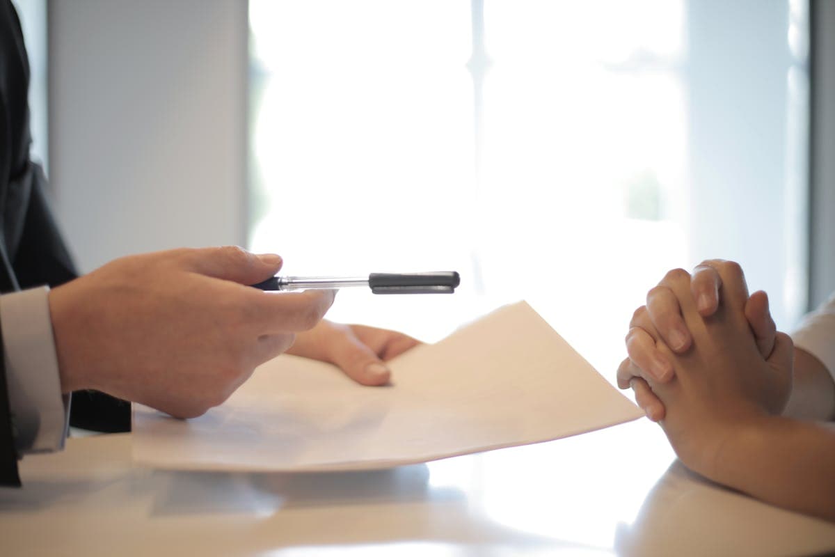 One person offering a pen and a contract across a table to another with folded hands
