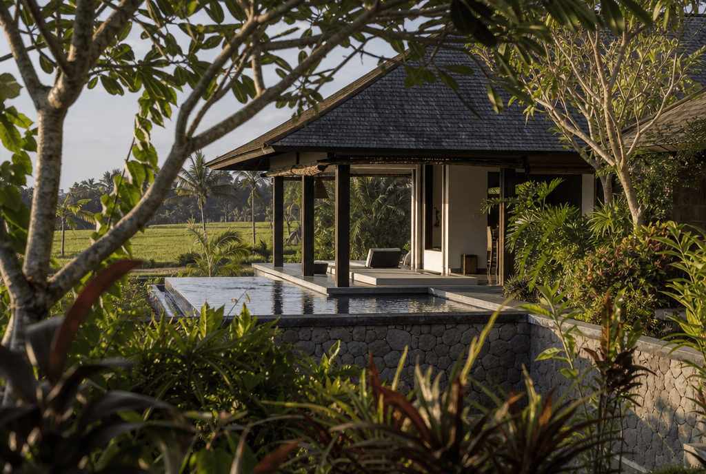 A Balinese villa with a pool and a stone boundary wall, seen from the garden, with rice fields behind it