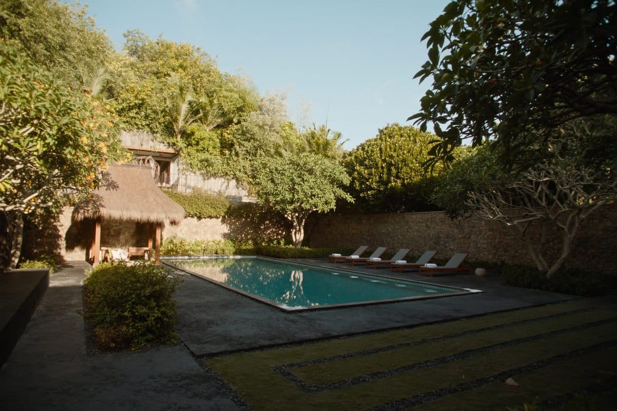 Well-kept Balinese villa garden with a lap pool, thatched bale and sun loungers beside a stone wall