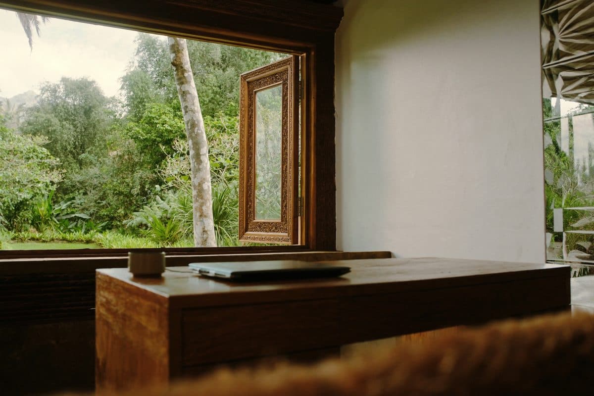 Laptop di atas meja kayu gelap di samping jendela yang menghadap rimbunnya hutan