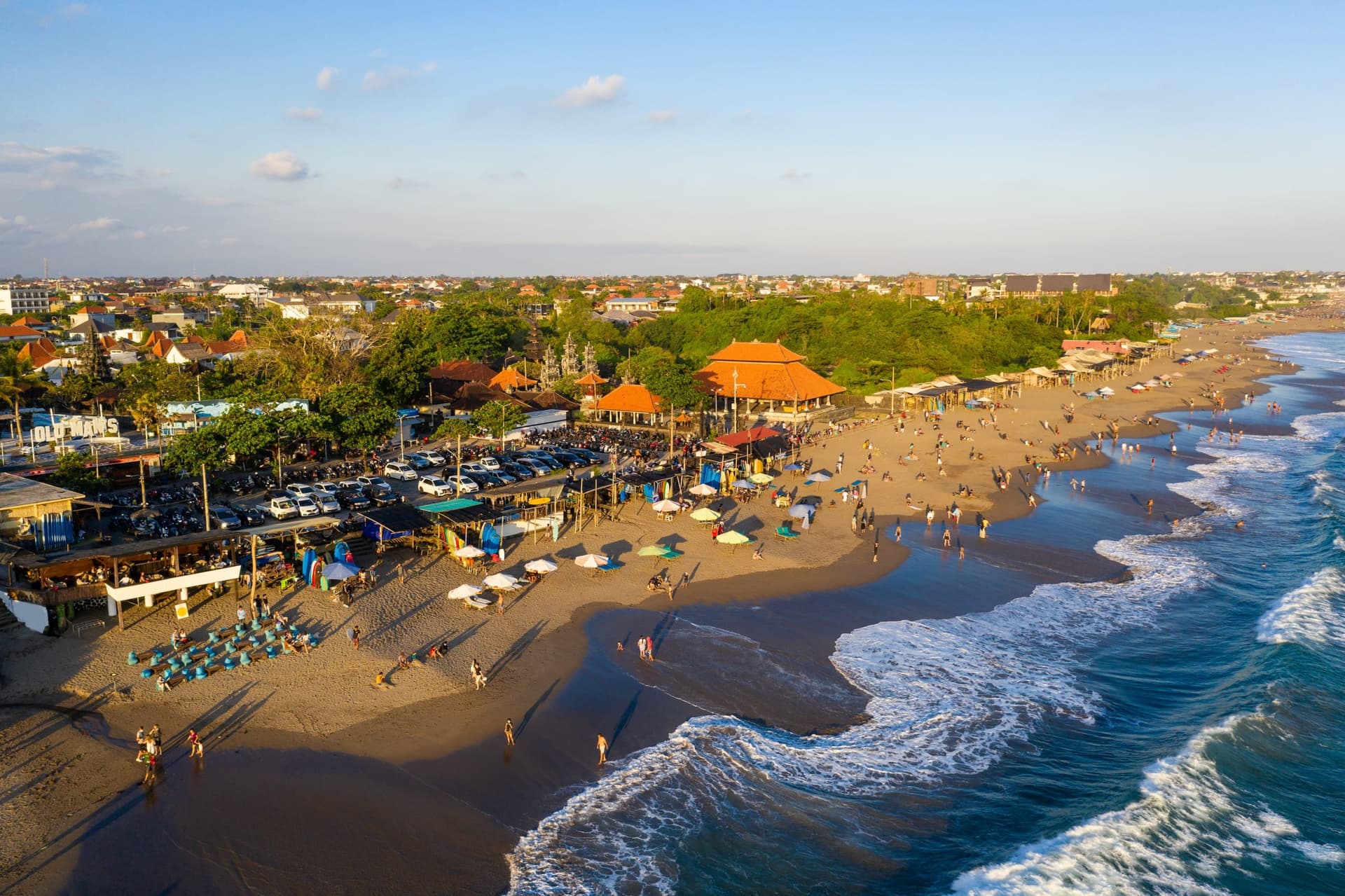 Aerial view of a busy Bali beach at golden hour, with beach clubs and warungs, rows of parked motorbikes, and surf rolling in.