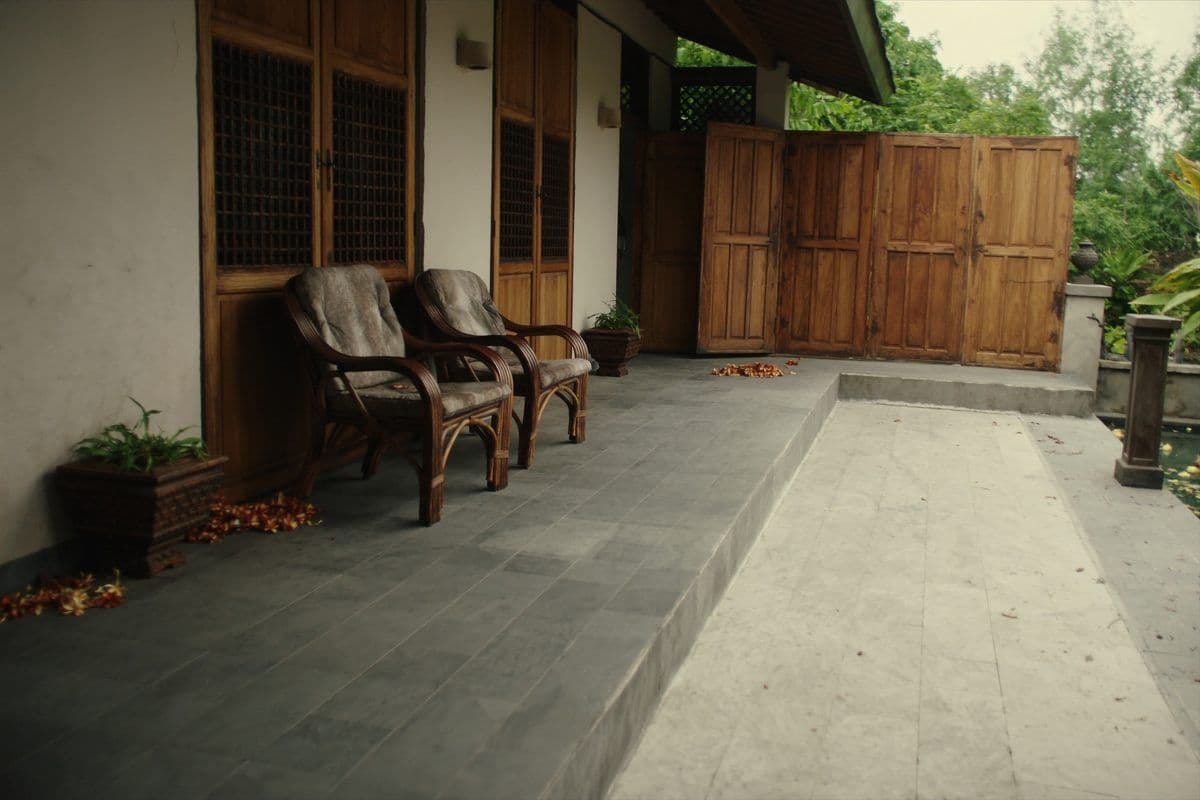 Empty Balinese villa terrace with rattan chairs, closed teak doors and fallen frangipani on the stone floor