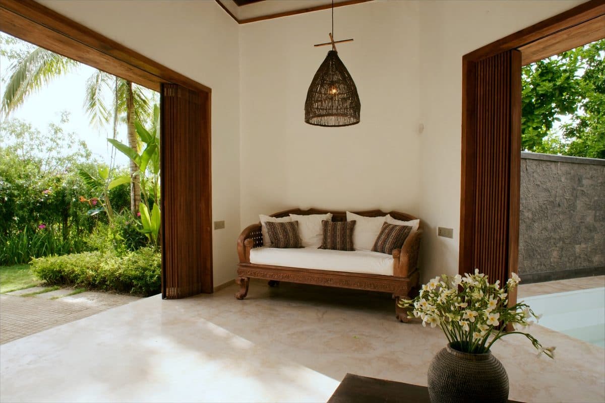 Carved teak daybed with ikat cushions in an open Balinese villa living space, folding doors onto a tropical garden