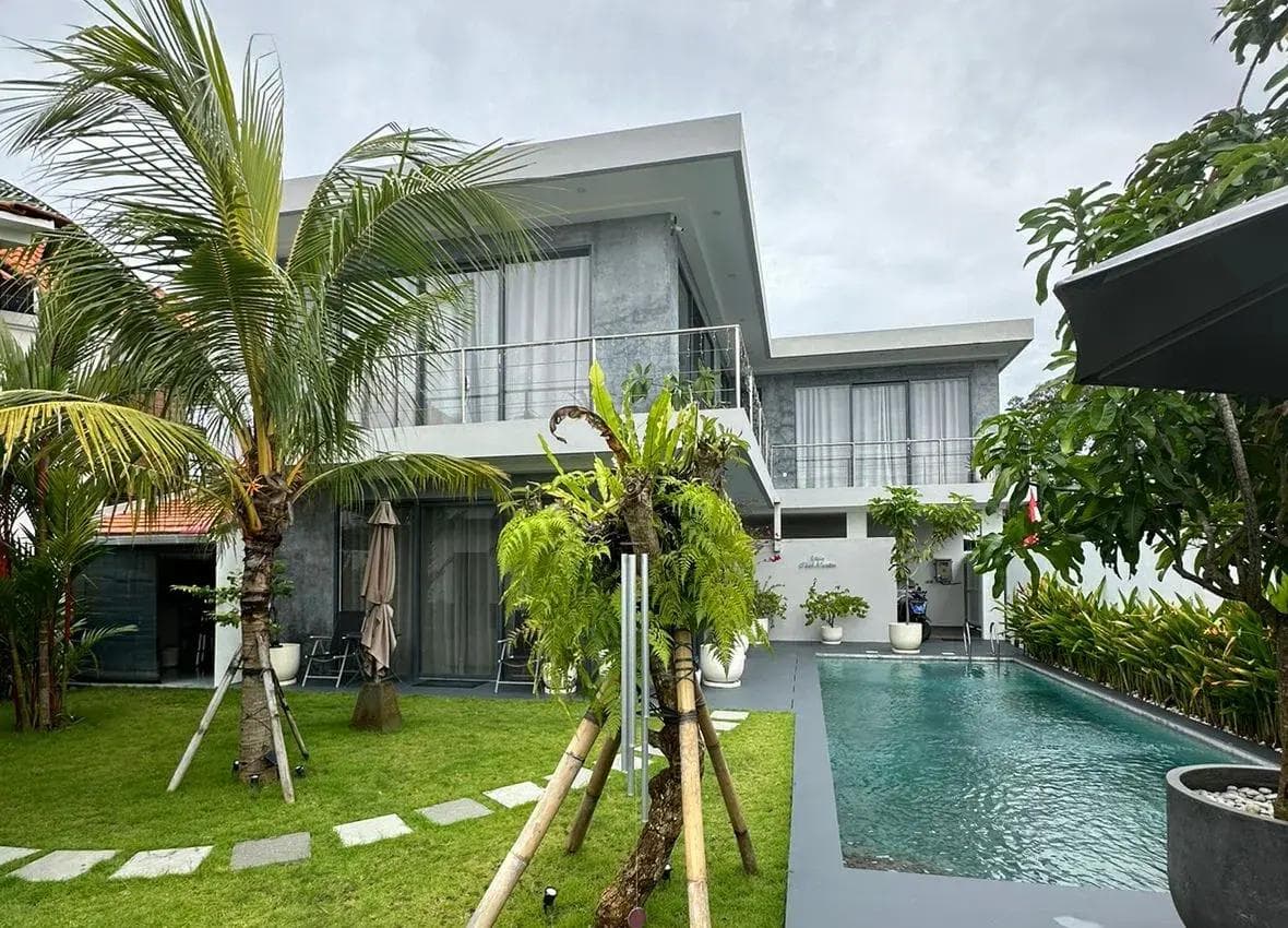 Modern two-storey villa with a grey concrete facade, a lap pool and young coconut palms staked in the lawn.