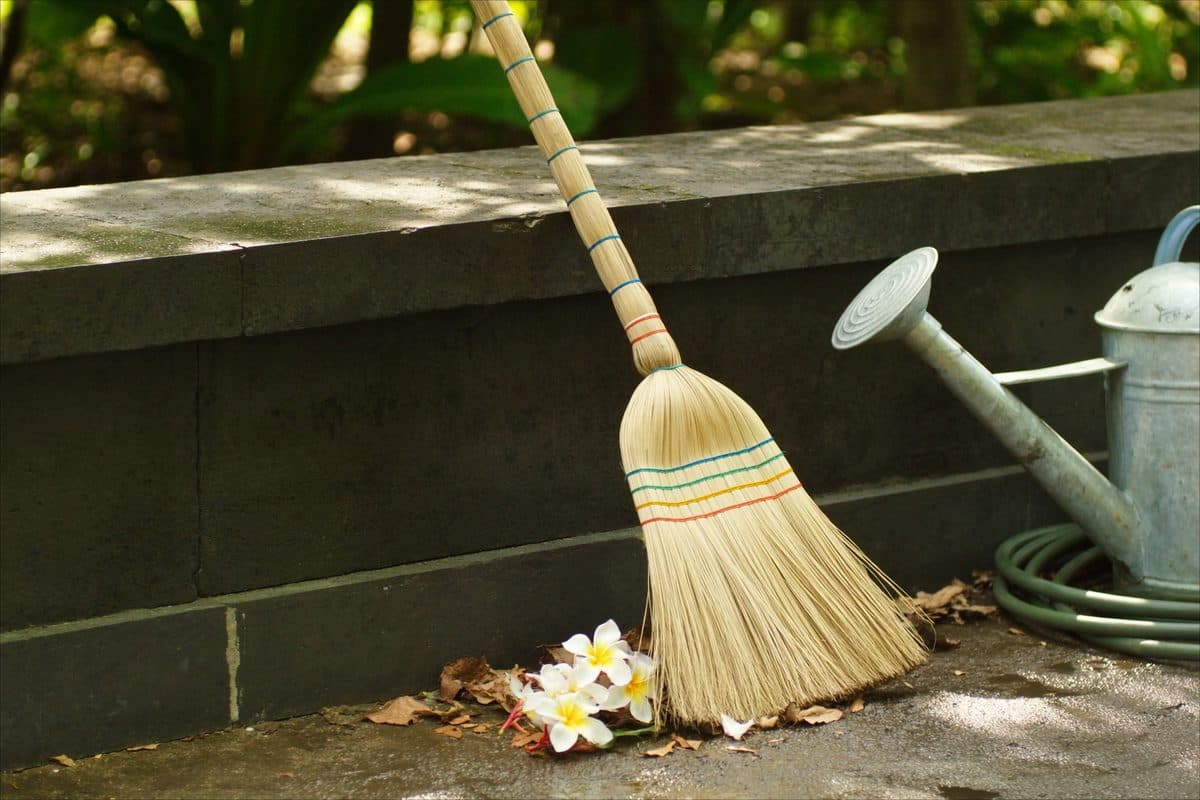 Indonesian palm-rib broom against a volcanic stone step, beside swept frangipani flowers, a watering can and a hose