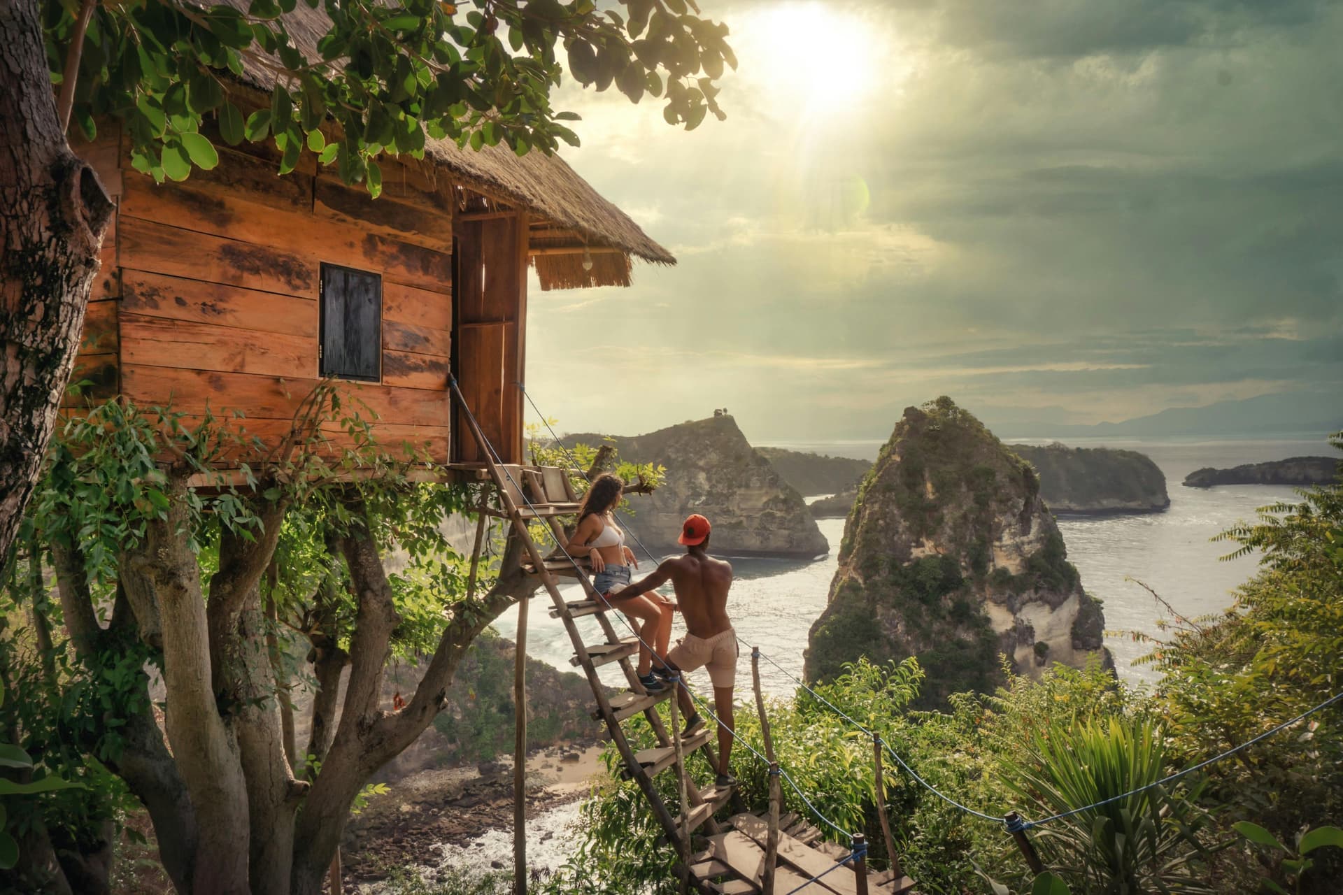 Two people on the ladder of a wooden cliffside treehouse, looking out over limestone sea stacks and a turquoise bay.